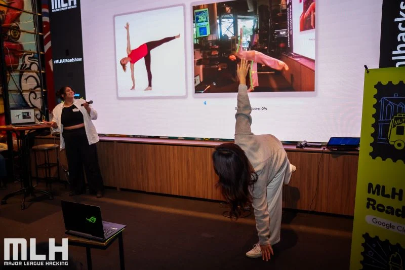 A presenter in a half-moon pose in front of the projected ZenPose screen, which shows the reference pose and her live camera view; her co-presenter holds a microphone to the left