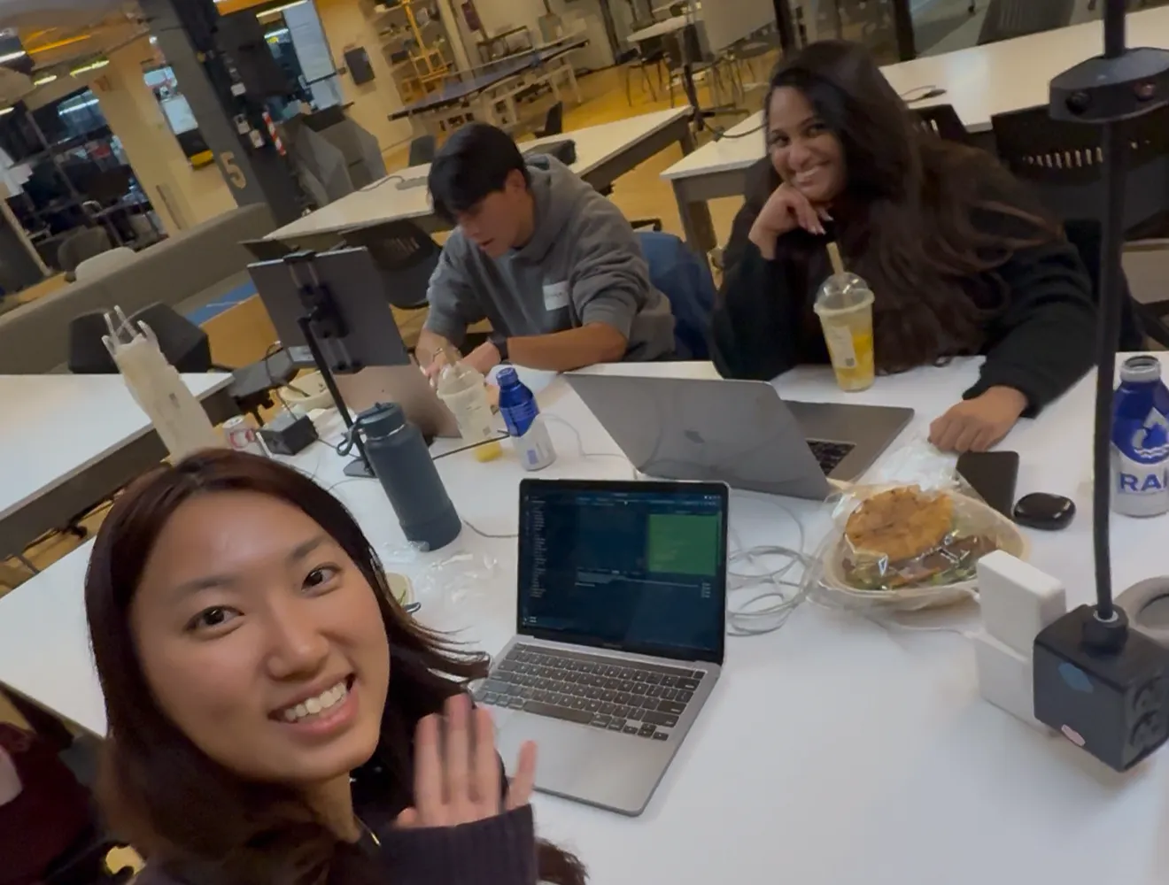 The team at their table at HackNYU: one teammate waving at the camera in a selfie, two more working behind her, with laptops and food on the table